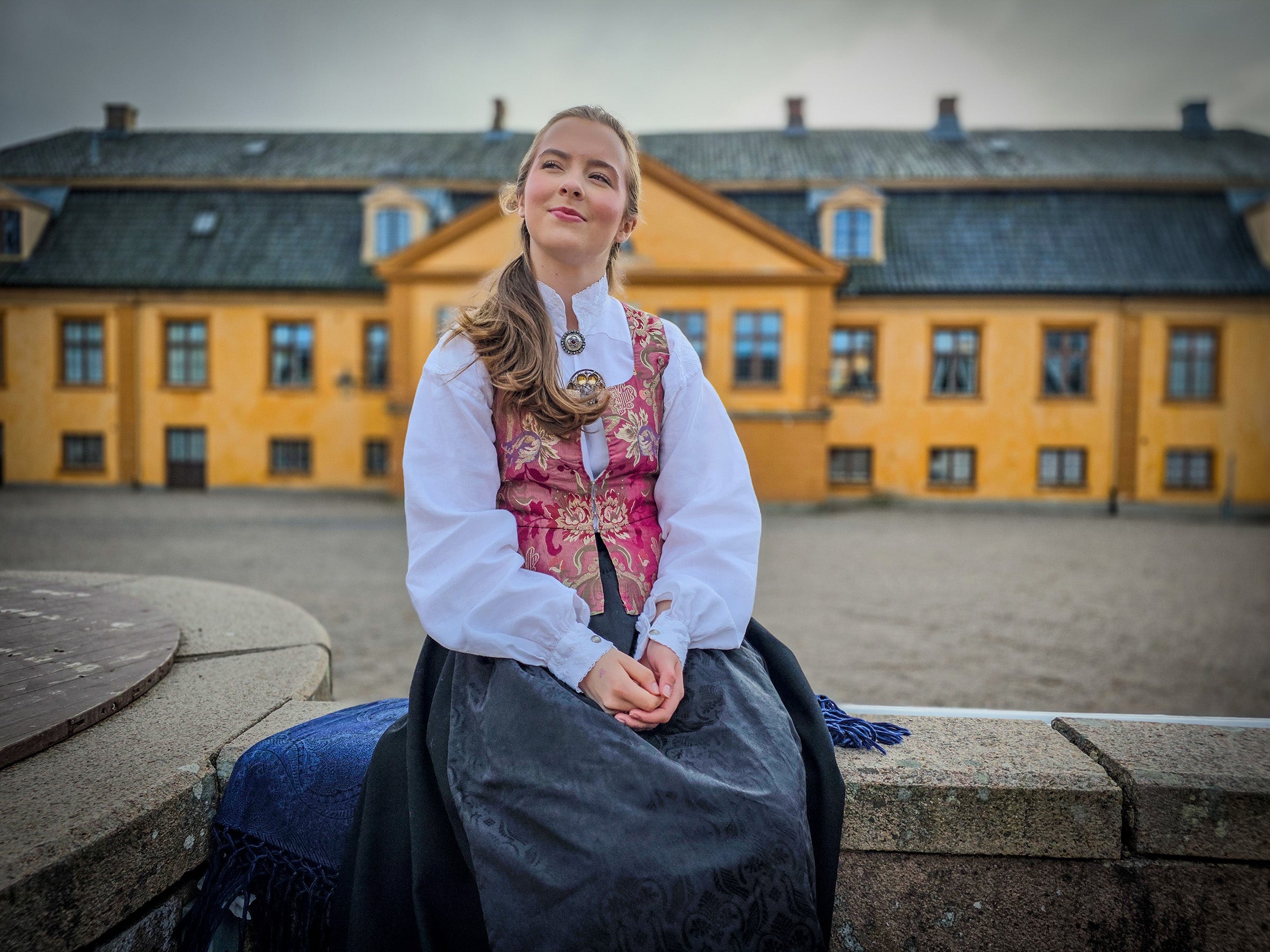 Konfirmant i Østerdalsbunad for dame med brokade vest som ser ut i luften på Bogstad Gård (hovedhuset i bakgrunnen)